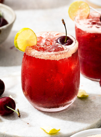 A vibrant red cherry limeade in a stemless wine glass, with a garnish of a cherry, a lime wedge, and a sparkling sugar rim.