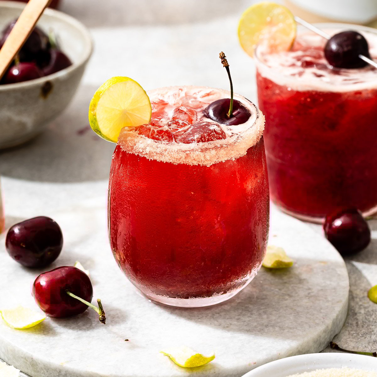 A vibrant red cherry limeade in a stemless wine glass, with a garnish of a cherry, a lime wedge, and a sparkling sugar rim.