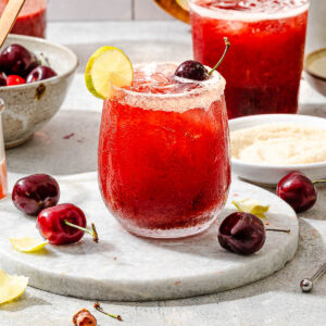 A vibrant red cherry limeade in a stemless wine glass, with a garnish of a cherry, a lime wedge, and a sparkling sugar rim.