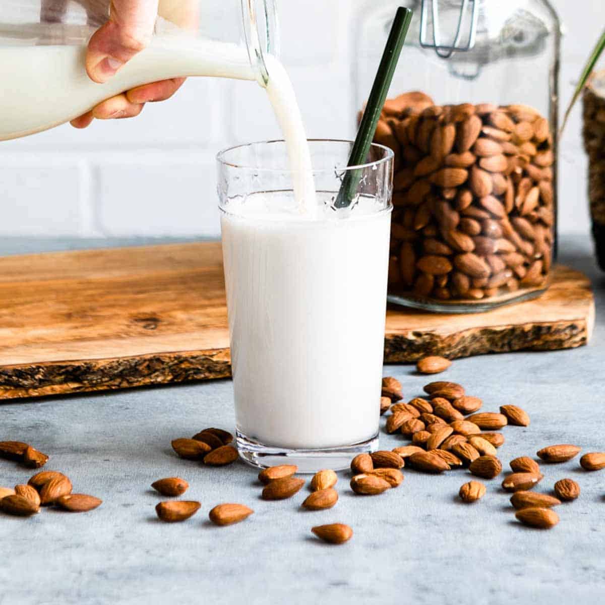 pouring. a glass of homemade milk into a cup with a green straw on a counter full of almonds.