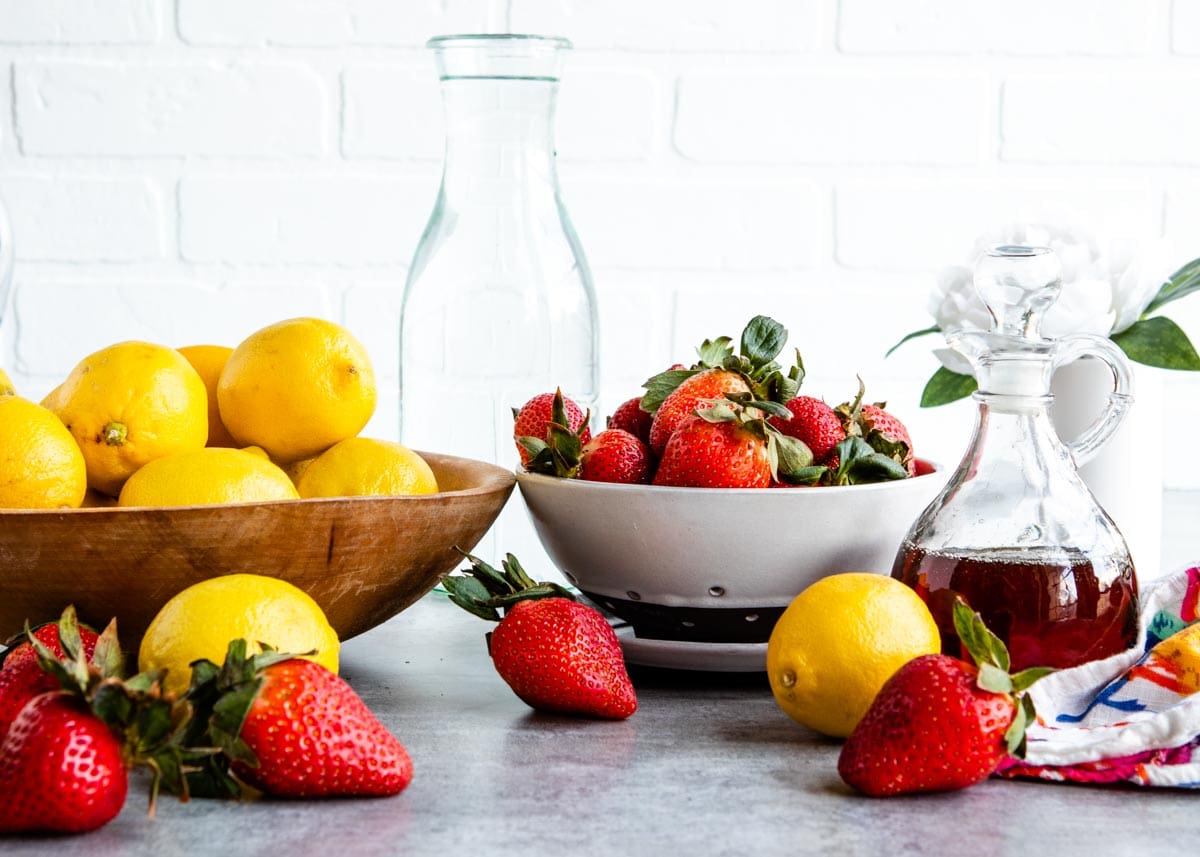bowl of lemons and bowl of fresh strawberries next to glass jar of maple syrup on a countertop.