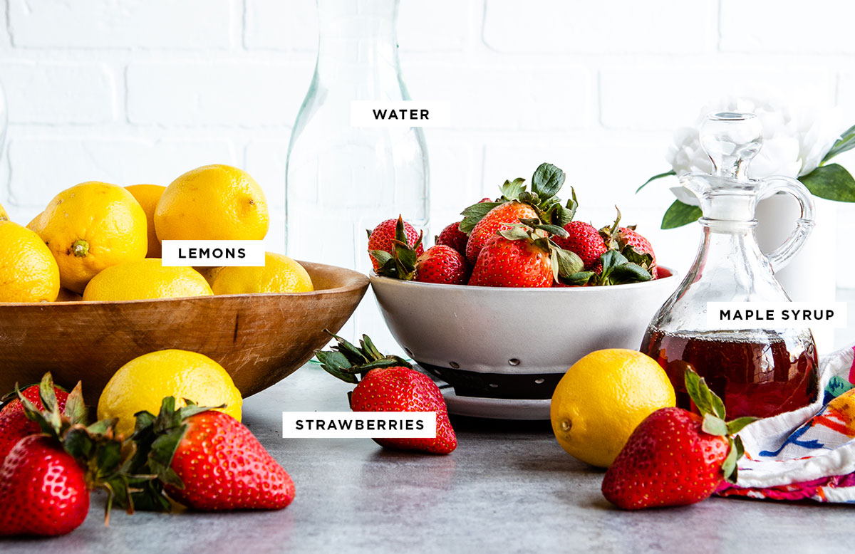 bowl of lemons and bowl of fresh strawberries next to glass jar of maple syrup on a countertop.