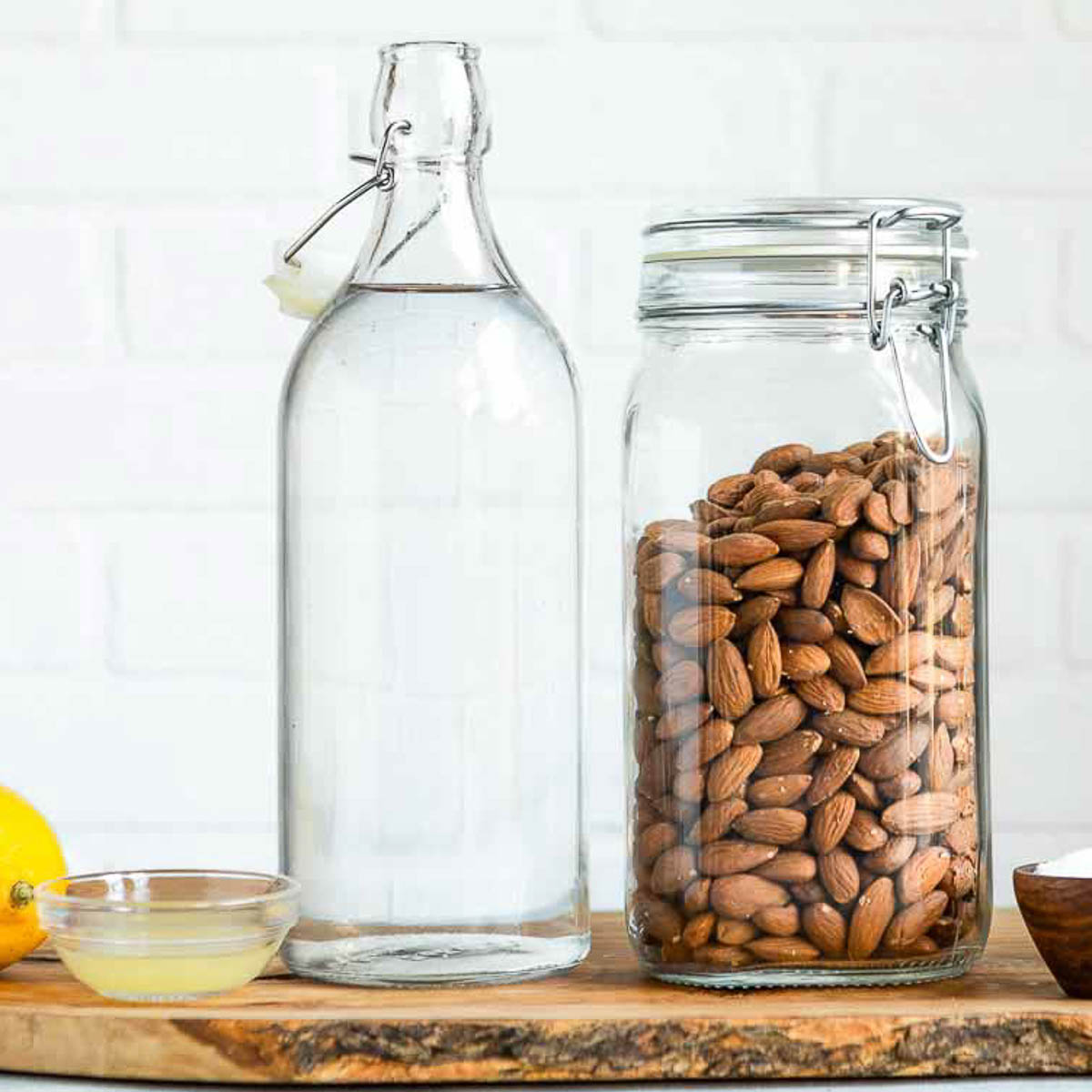 A glass bottle of water and a jar of almonds on a wooden cutting board.