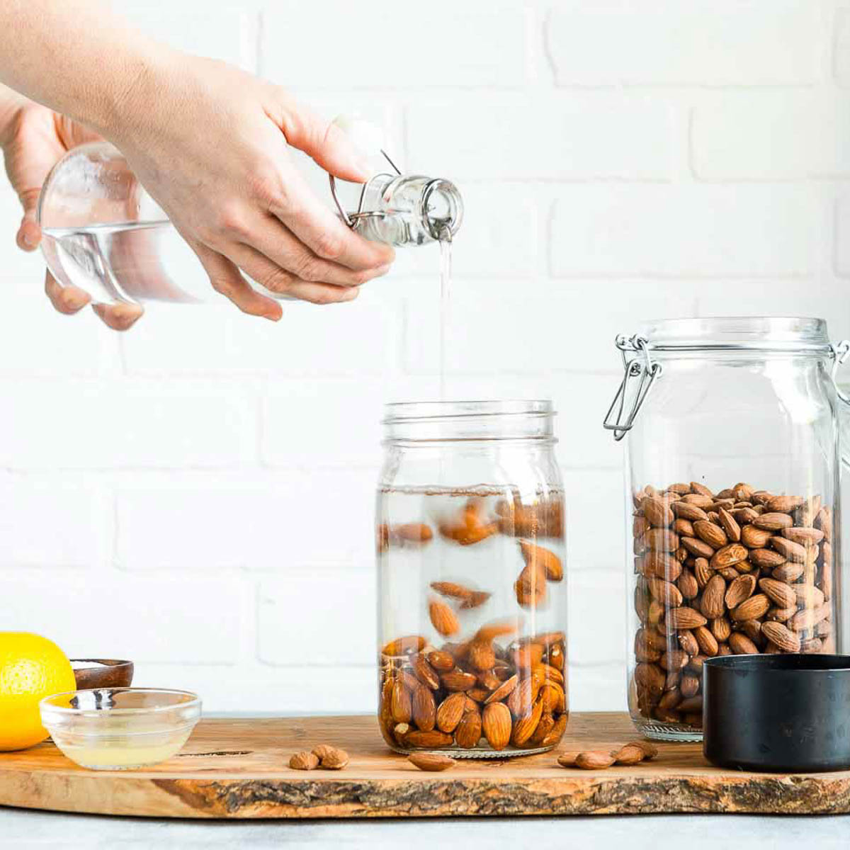 A person is adding water to a jar of almonds, with a lemon and other ingredients visible.