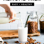 Homemade almond milk being poured into a glass surrounded by scattering of raw almonds.