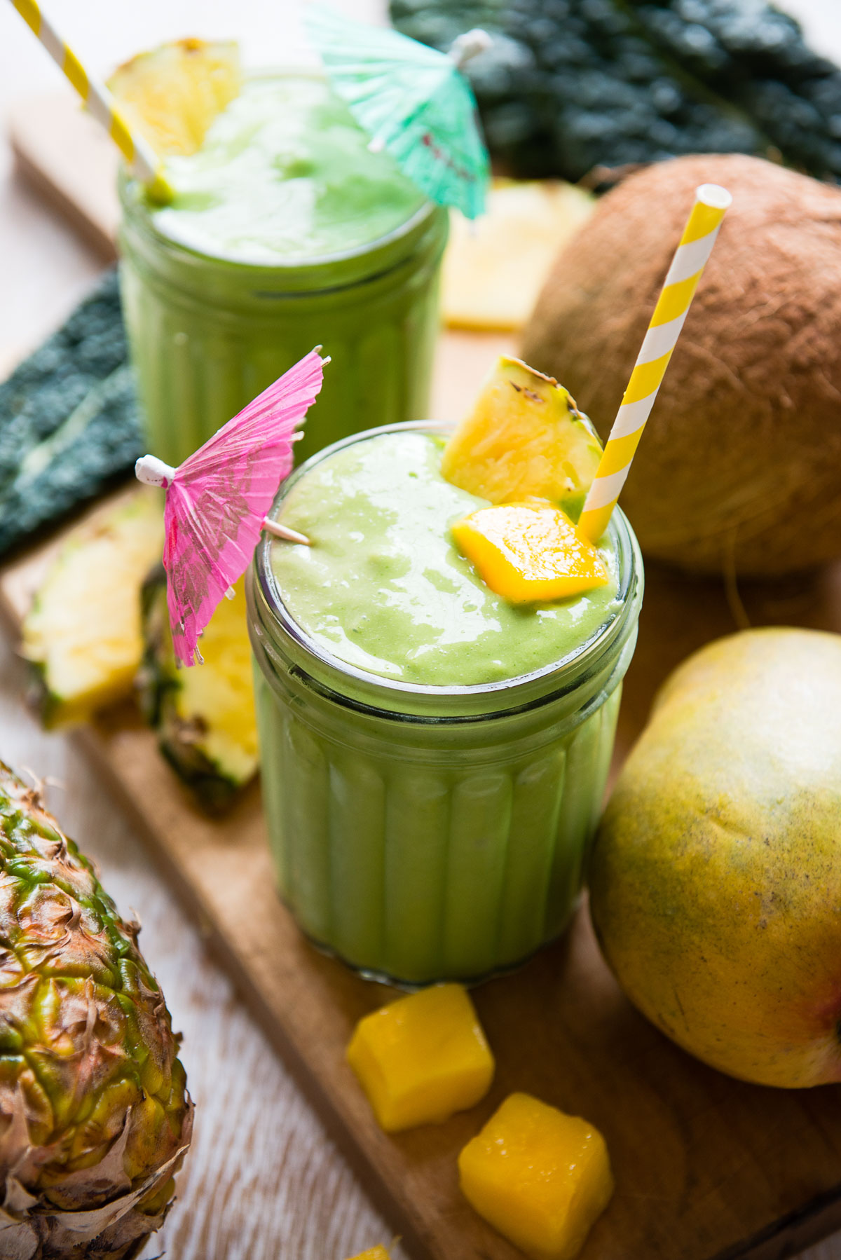 Two kale mango smoothies in glasses with tropical umbrellas, surrounded by fresh ingredients.