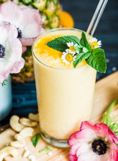 A vibrant yellow pineapple smoothie, garnished with fresh mint, and surrounded by pink flowers