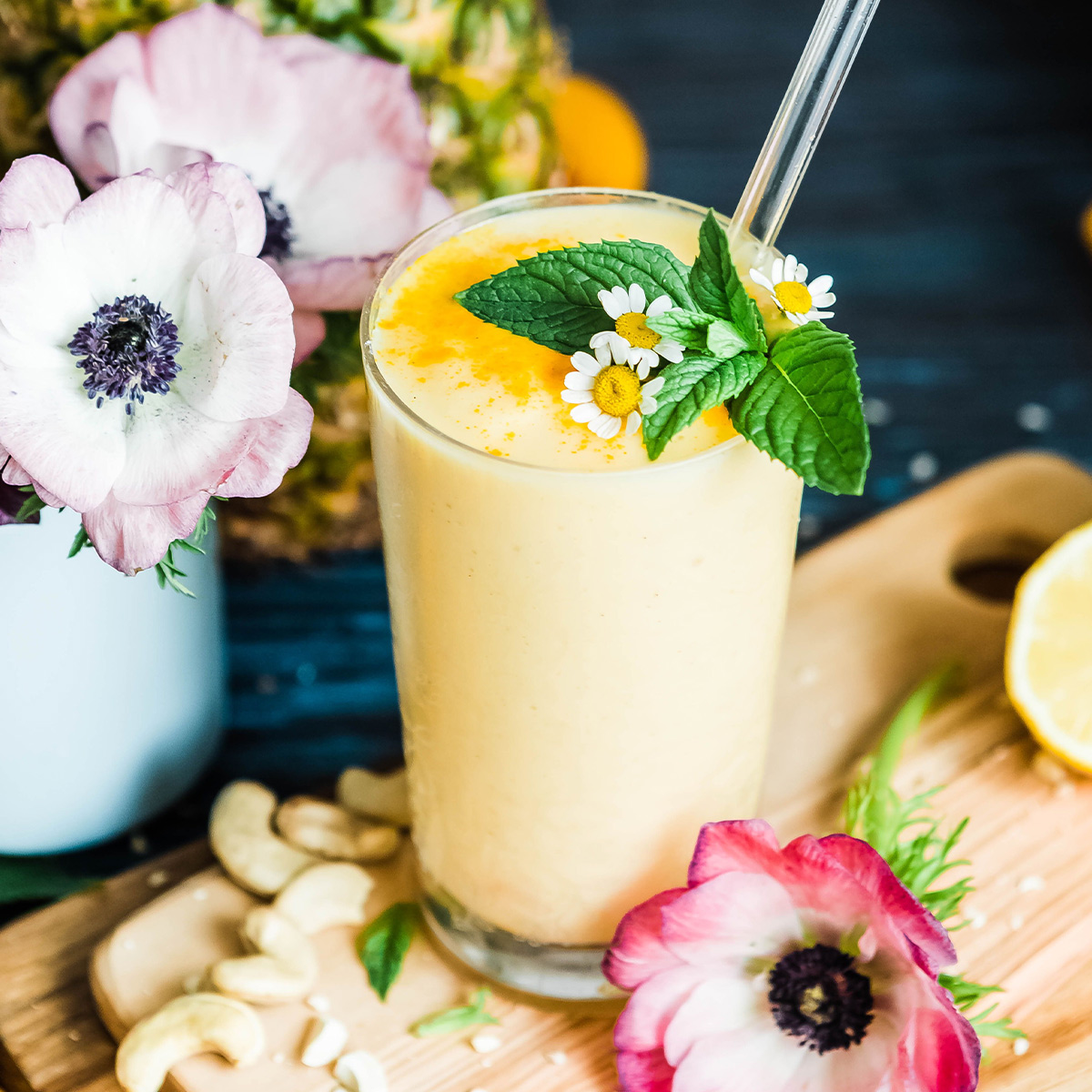 A vibrant yellow pineapple smoothie, garnished with fresh mint, and surrounded by pink flowers