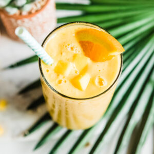 Top-down view of a mango smoothie garnished with an orange slice, mango chunks, and a paper straw.