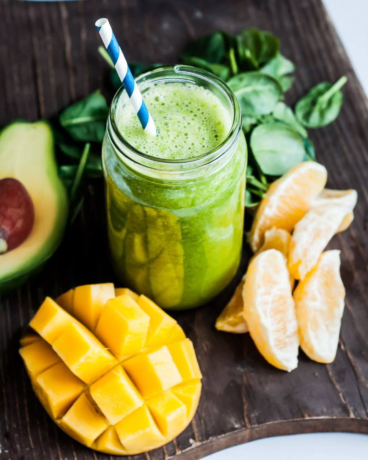 Mango spinach smoothie in a Mason jar surrounded by orange slices, spinach, mango and Avocado.