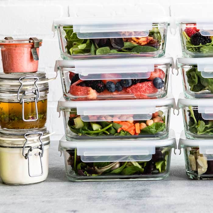 8 glass containers of meal prep salads next to 3 glass jars of salad dressing.