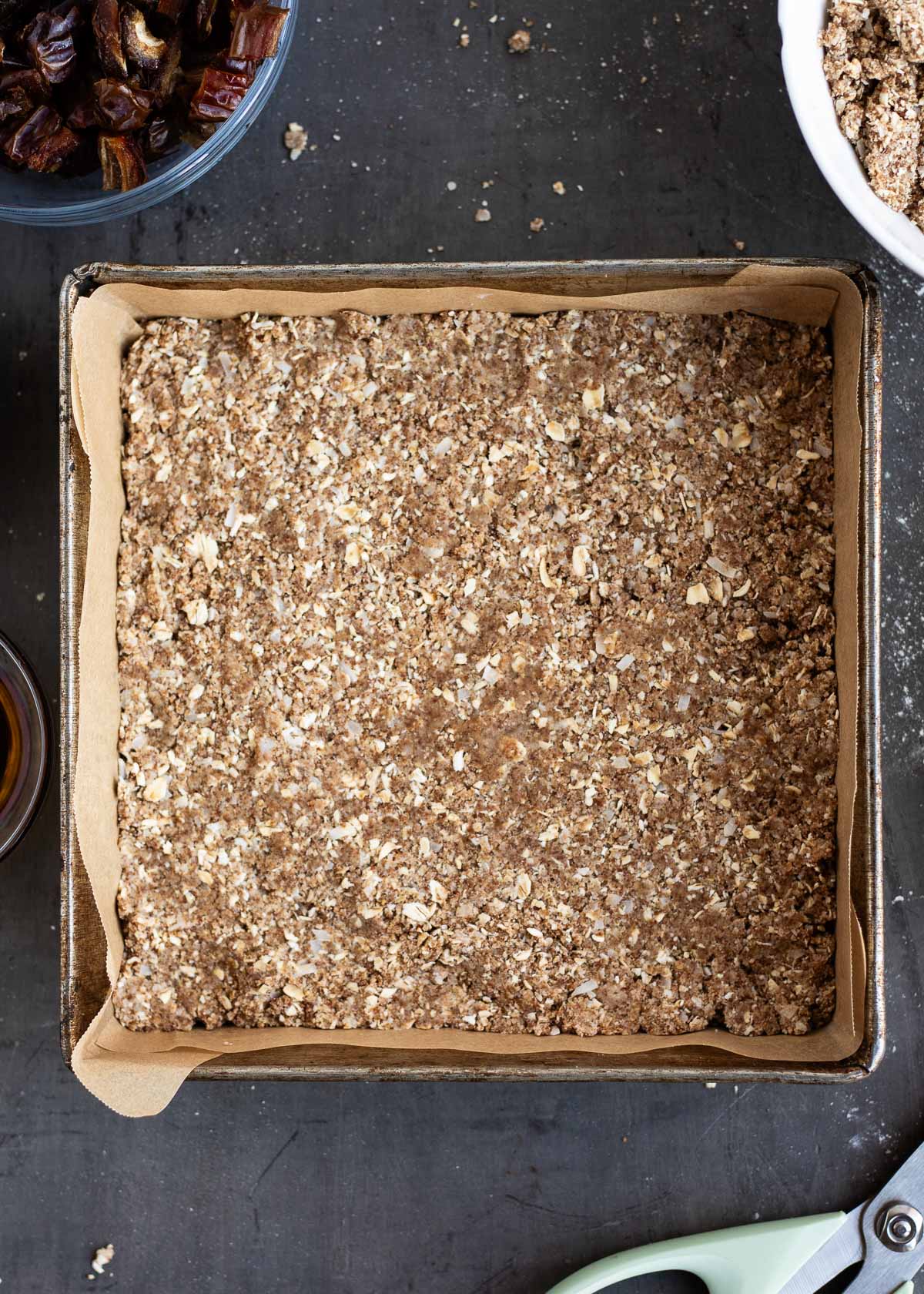 A baking pan lined with parchment paper and filled with a flattened, crumbly base layer