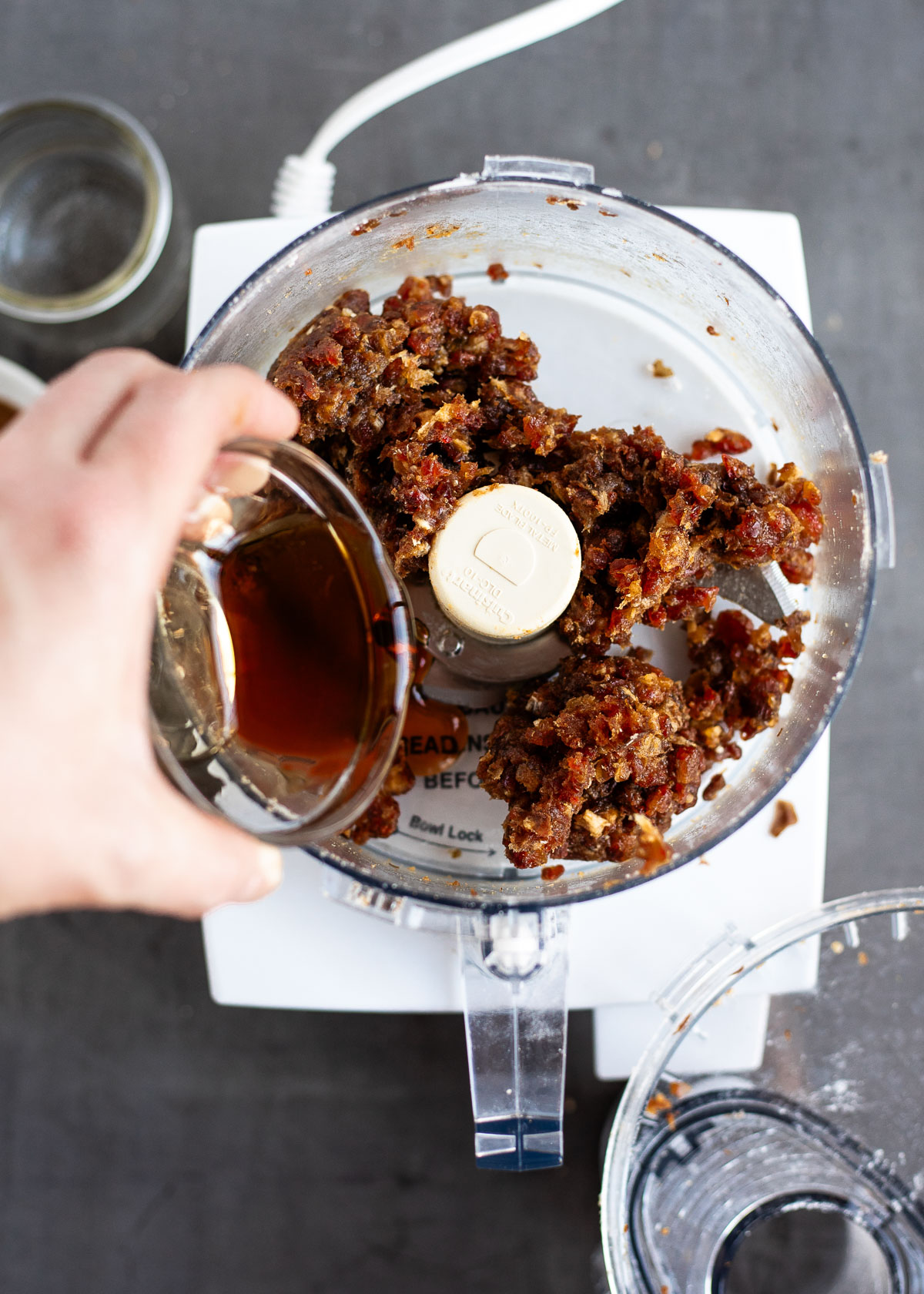 A person drizzling maple syrup into a food processor filled with blended dates