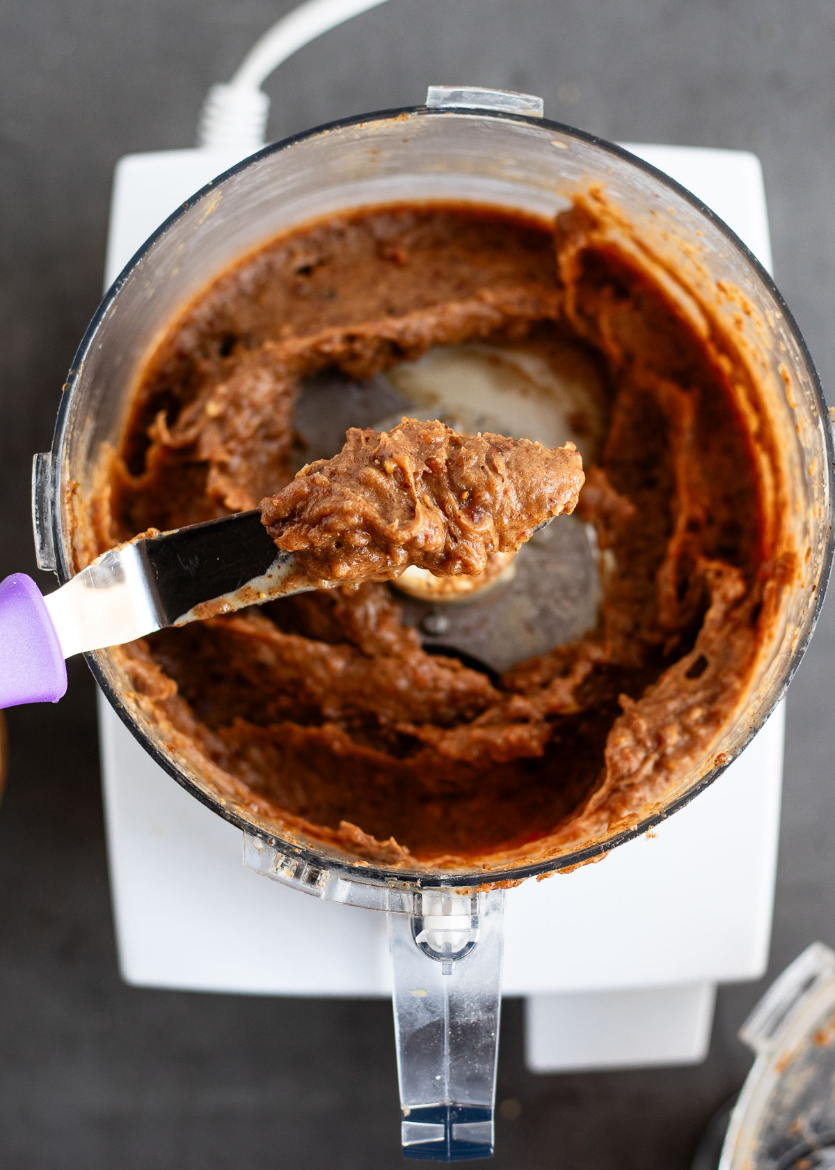 A knife scraping out the thick, sticky date mixture from a food processor