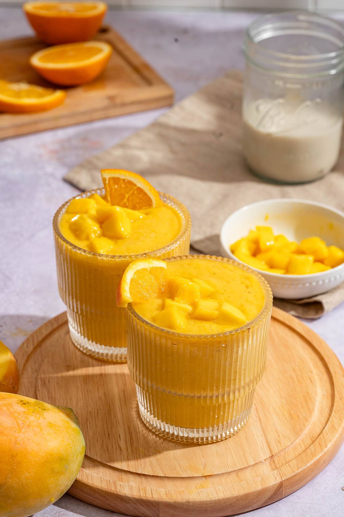 Two glasses of orange mango smoothie on a round wooden board with orange slices, a mango half and mango chunks.
