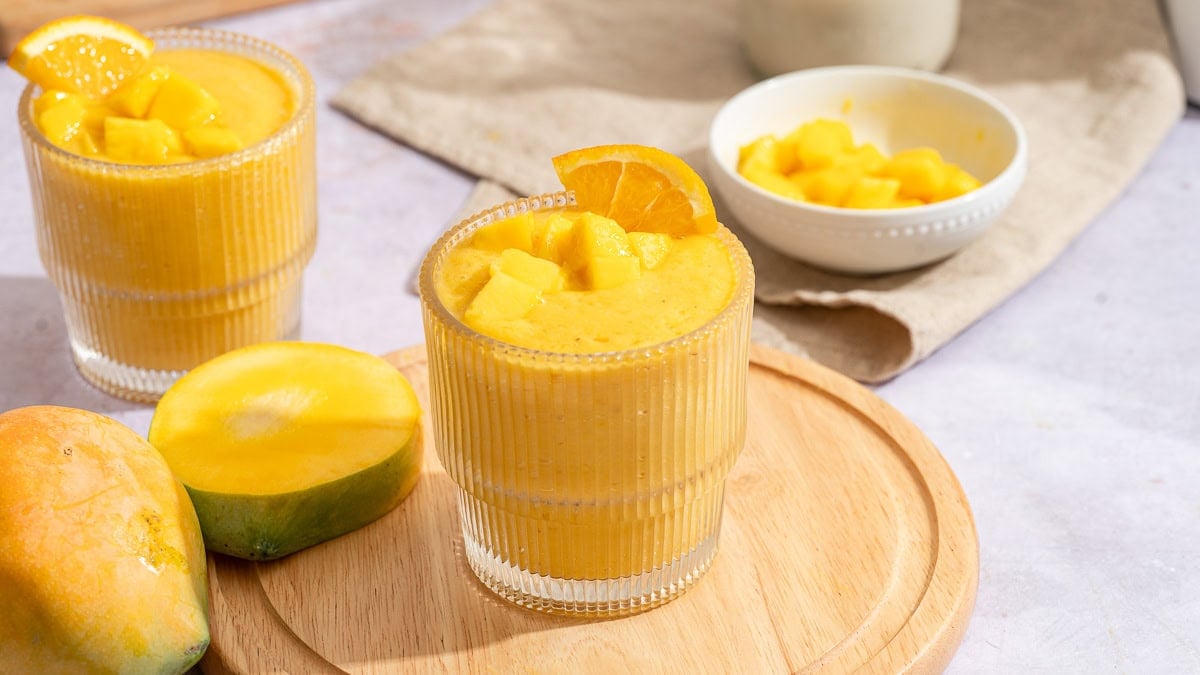 Orange mango smoothie in a ribbed glass topped with an orange slice, with a mango half and a bowl of mango chunks nearby.