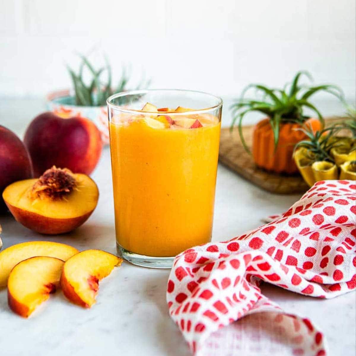 glass of peach smoothie topped with fresh peaches, on a white countertop with whole and sliced peaches.