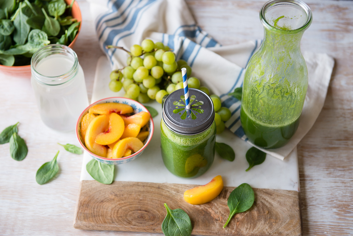 green smoothie in a glass mug next to a liter of green smoothie and bowls of grapes, peaches, coconut water and spinach.