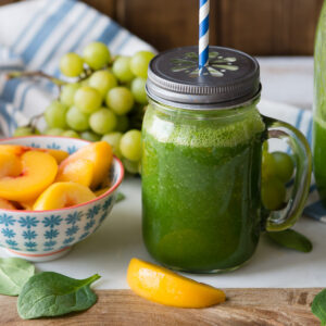 Peach spinach smoothie in a glass Mason jar with a lid and straw next to fresh ingredients