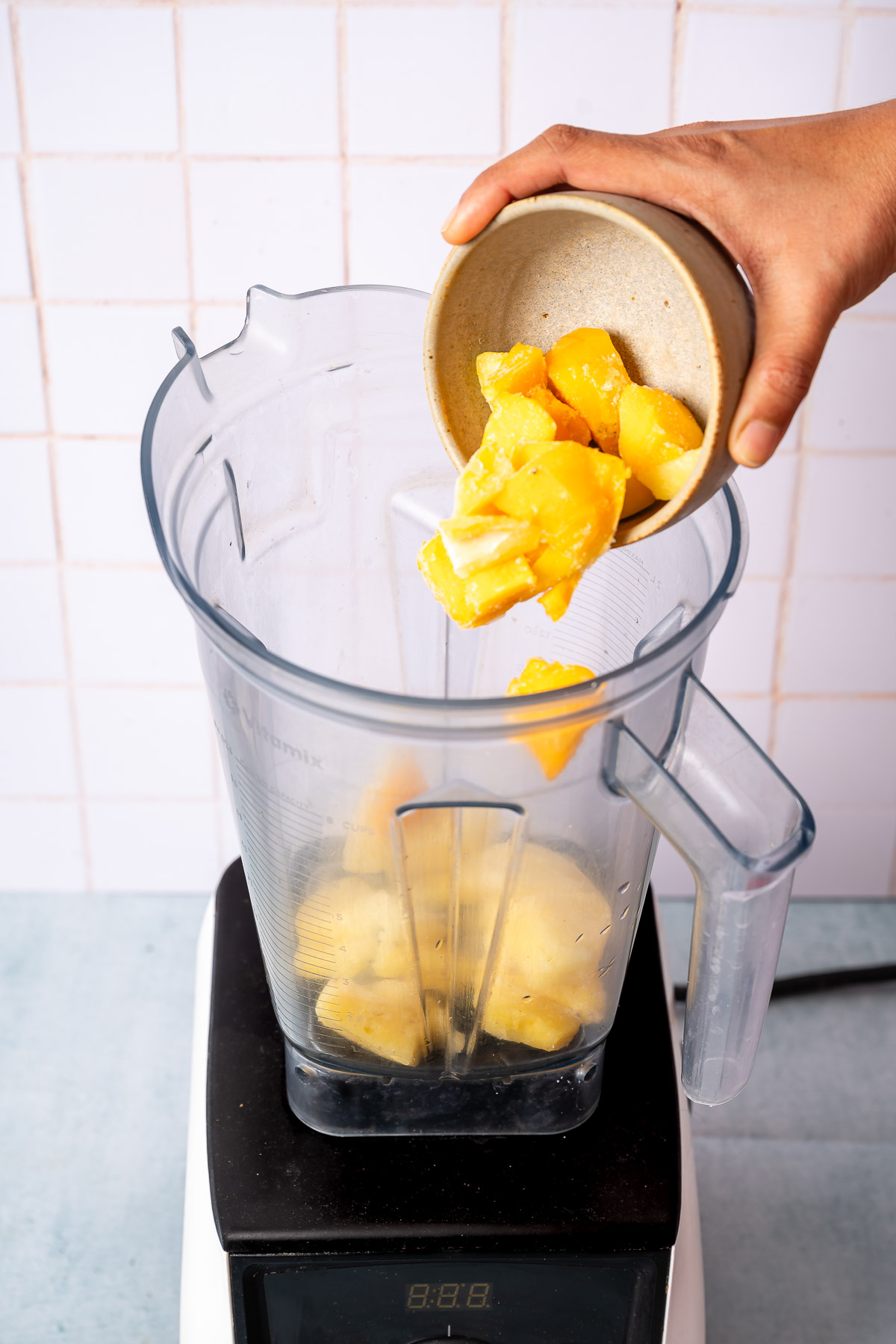 A person adding frozen pineapple and mango chunks into a blender