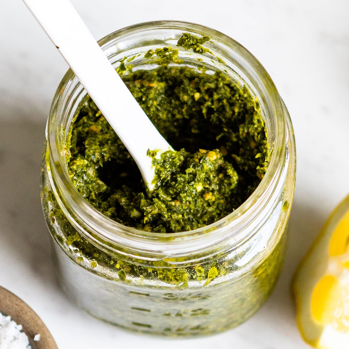 jar of pistachio pesto with ingredients used to make it sitting all around: lemons, pistachios, salt, herbs