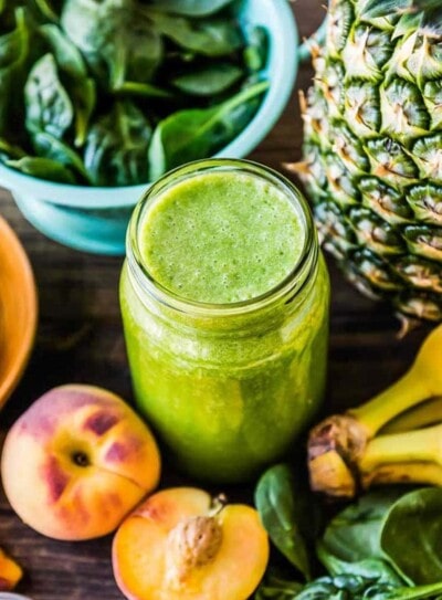 glass jar full of green smoothie, surrounded by fresh produce including pineapple, bananas, peaches and spinach.