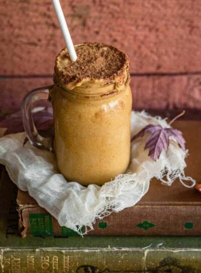 maple pumpkin fall smoothie in a glass mug with a paper straw on top of cheese cloth and a stack of antique books.