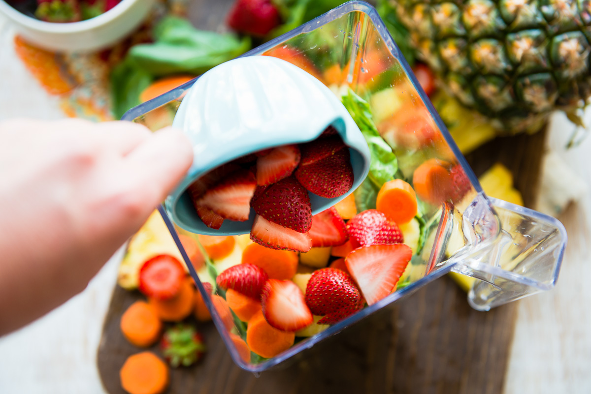 Hand pouring fresh, sliced strawberries from a blue measuring cup into a blender container.