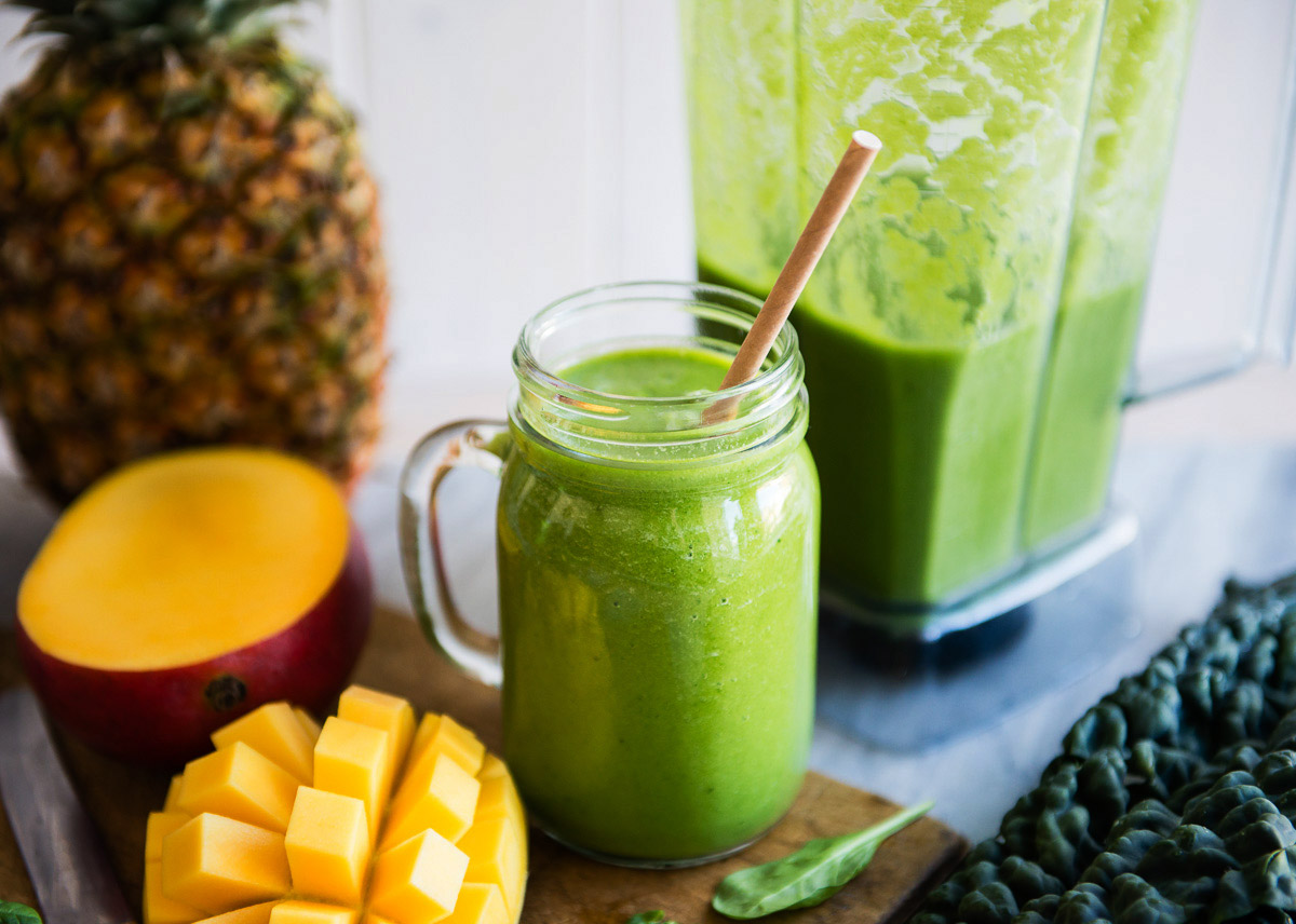 Detox smoothie in a mason jar with a paper straw surrounded by fresh mango and pineapple.