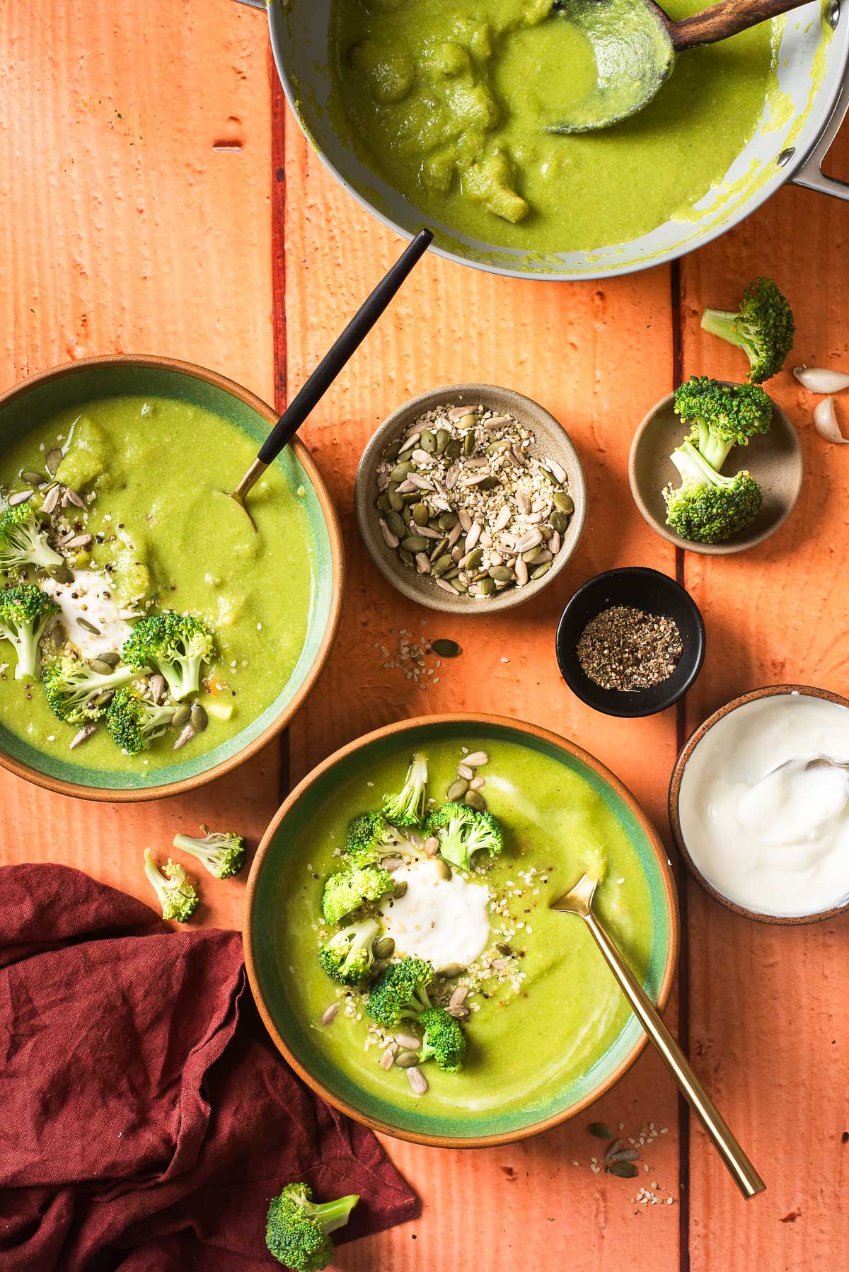 Two bowls of ready-to-eat vegan broccoli soup, with toppings in separate bowls next to the soup pot.