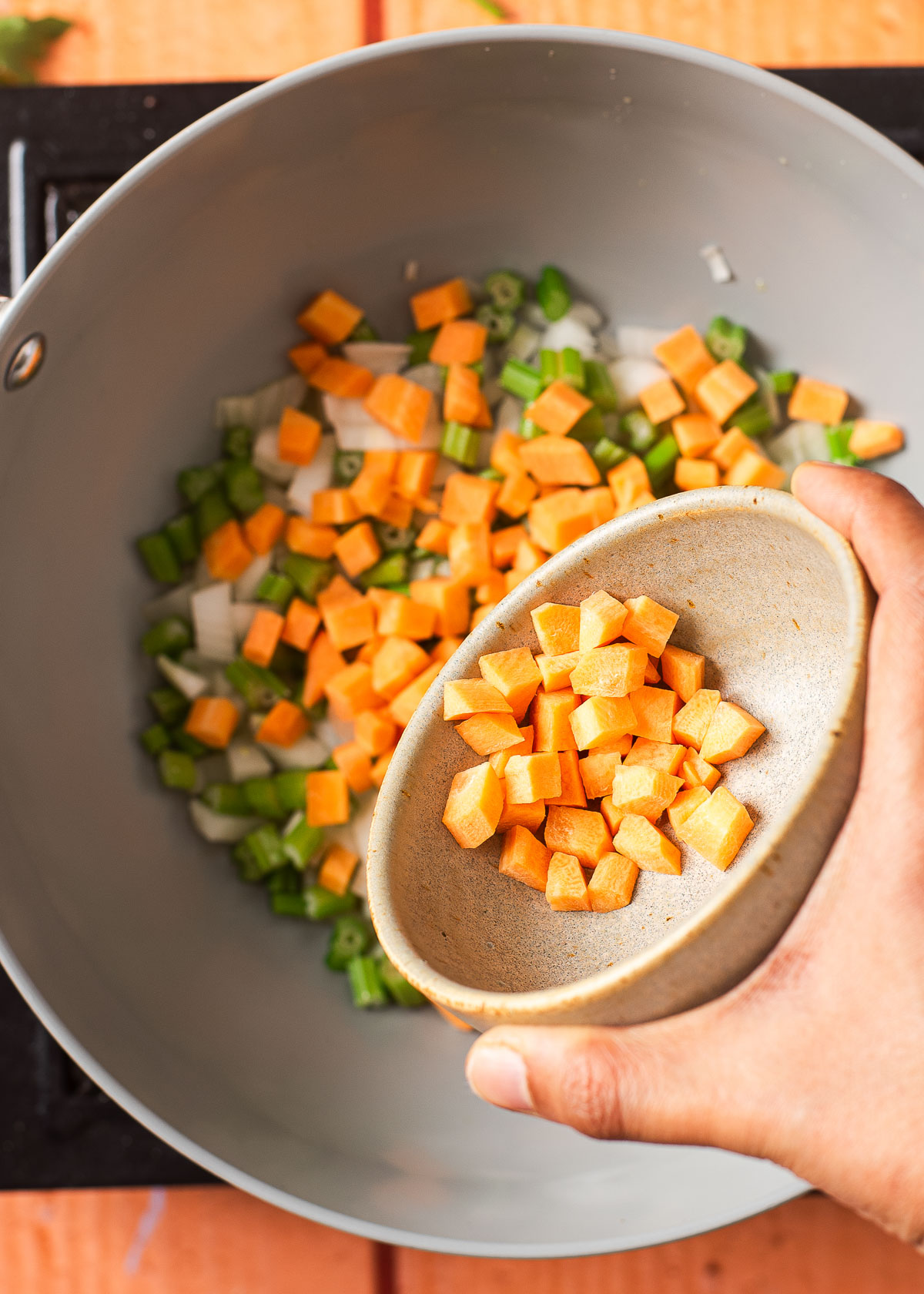 A hand pouring a small bowl of diced carrots into a pot with onions and celery