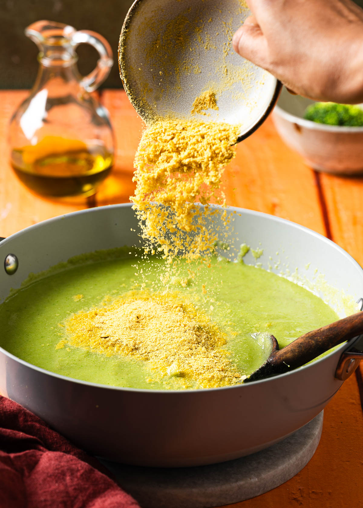 A hand pouring a bowl of nutritional yeast into a pot with vegan broccoli soup.