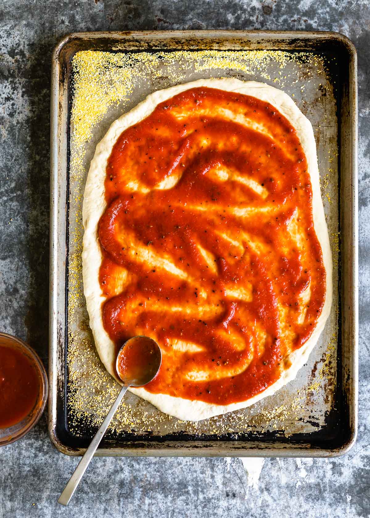 A spoon spreading pizza sauce over pizza crust