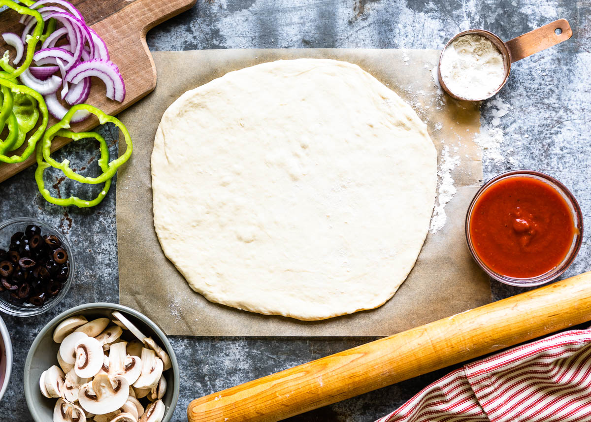Unbaked pizza dough rolled out surrounded by pizza ingredients
