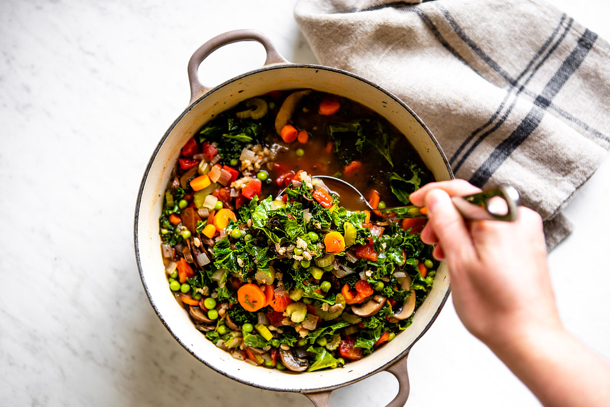 Lady stirring a big white pot of vegetable barley soup with an off white kitchen town to the side.