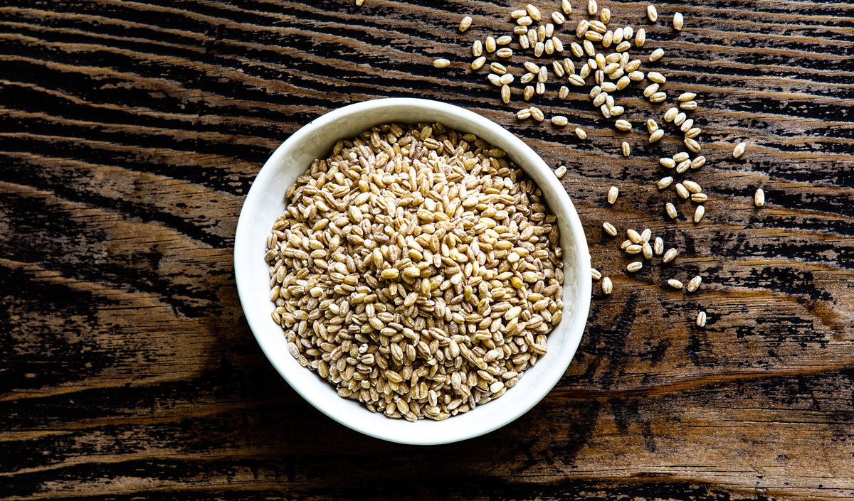 Barley close up shot in a white bowl with a few grains sprinkled onto a wood surface.