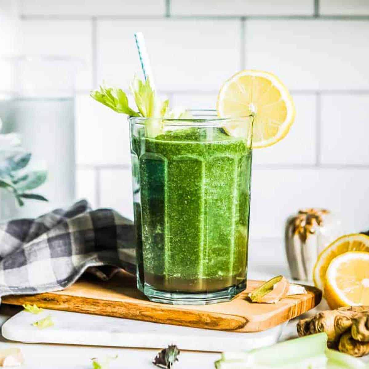 green smoothie in a glass cup with a straw and lemon garnish on a wooden cutting board.