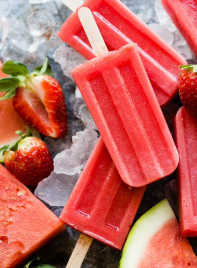 Fresh watermelon popsicles sitting on ice with fresh strawberries and watermelon slices.