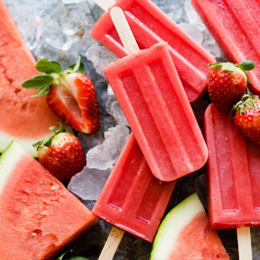 Fresh watermelon popsicles sitting on ice with fresh strawberries and watermelon slices.