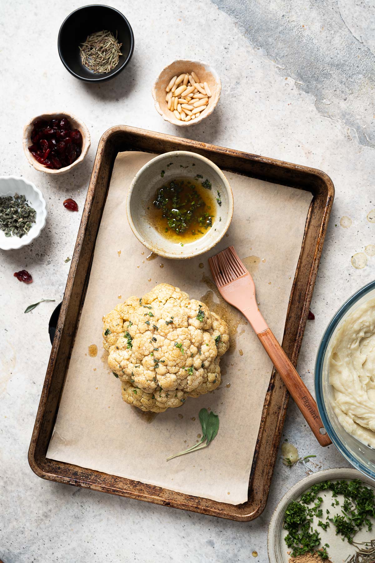 A bowl of oil and herbs next to a head of cauliflower, illustrating the process of brushing the mixture on top.