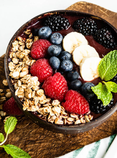 acai smoothie bowl in a coconut bowl on a wooden plank topped with granola, fresh berries, banana and mint leaves.