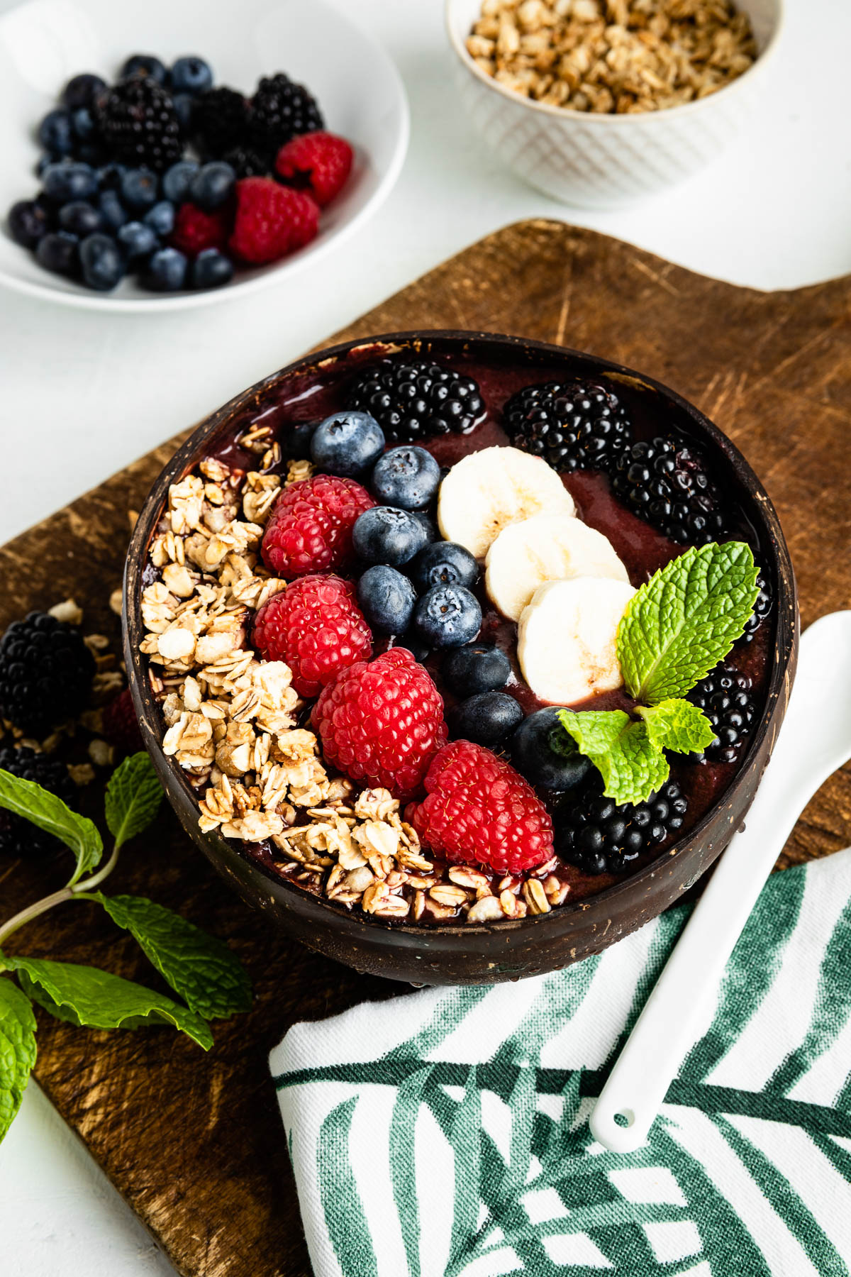 acai bowl in a coconut bowl on a wooden plank topped with granola, fresh berries, banana and mint leaves.