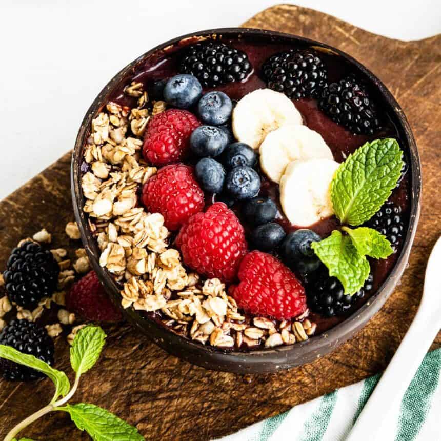 acai smoothie bowl in a coconut bowl on a wooden plank topped with granola, fresh berries, banana and mint leaves.