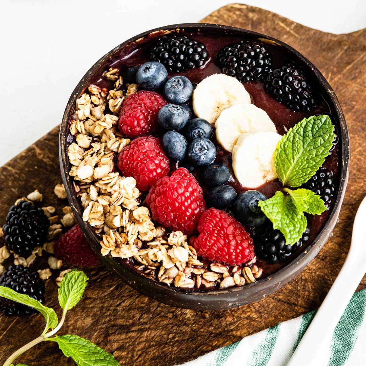 acai smoothie bowl in a coconut bowl on a wooden plank topped with granola, fresh berries, banana and mint leaves.