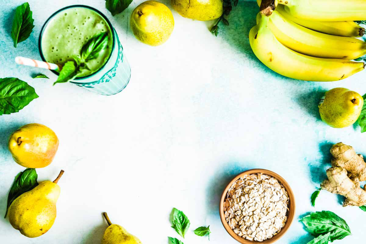 Bowl of oatmeal, fresh ginger root, bananas, pears and other acid reflux smoothie ingredients on teal counter next to a smoothie.