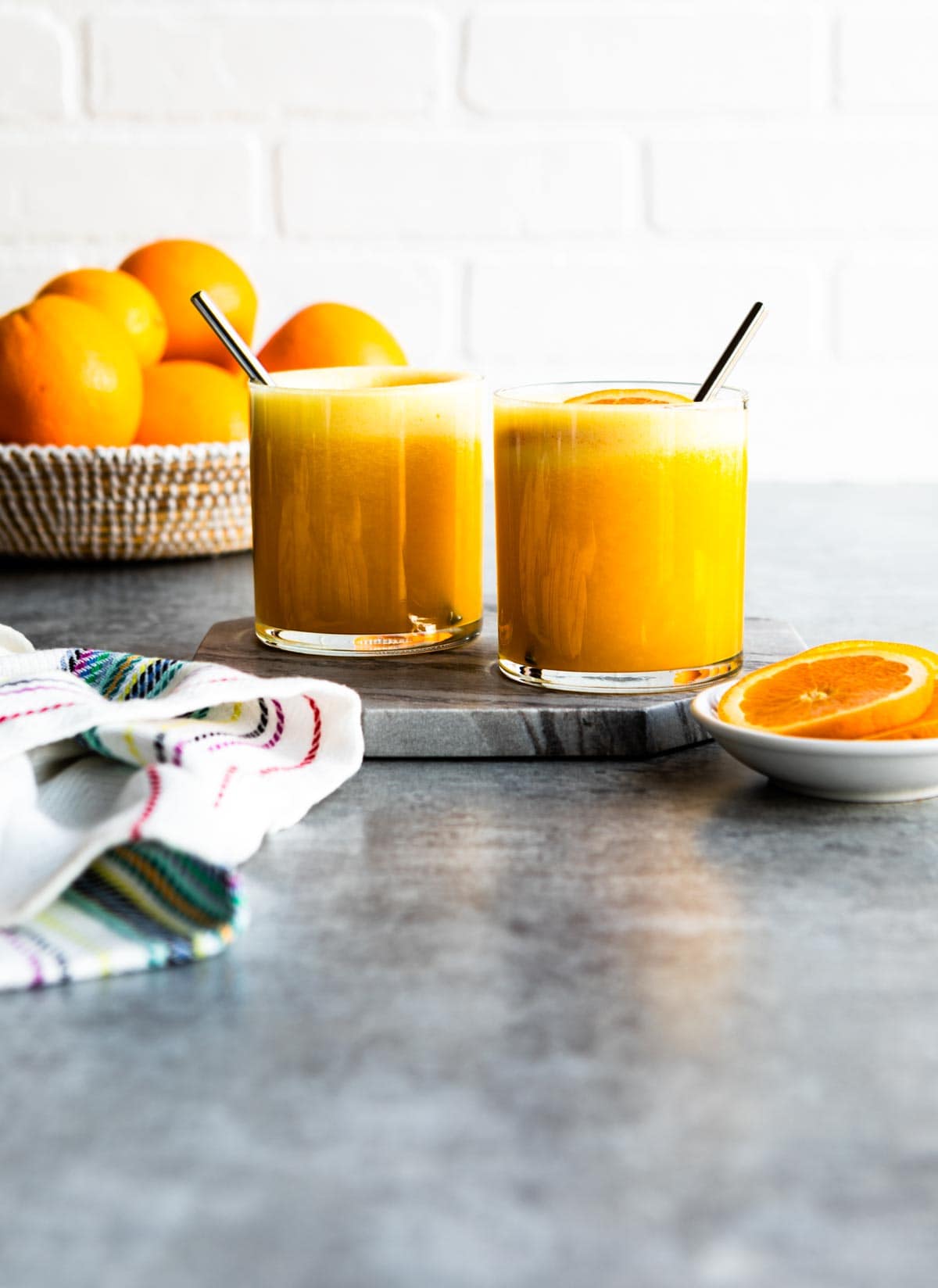 2 short glasses of orange beverage on a marble tray next to a basket of oranges.