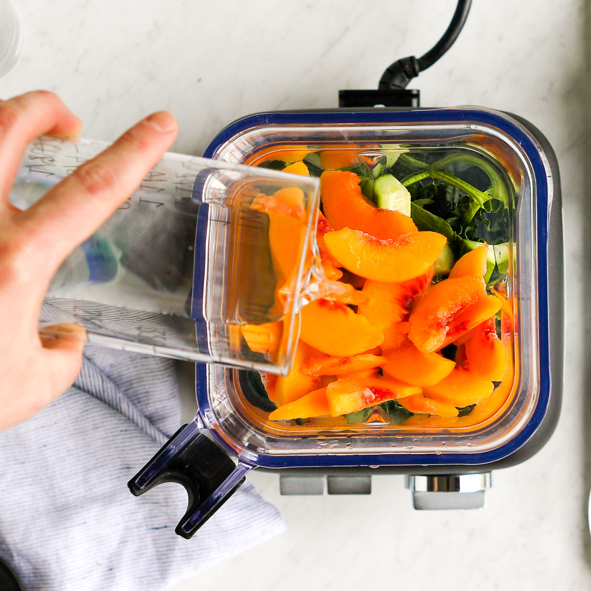Pouring water into blender with frozen peaches and kale.