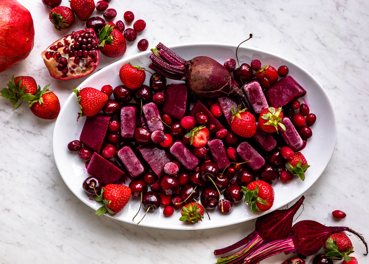 Smoothie cubes on a white plate scattered amongst fresh berries, cherries, beets, and other red and purple fruits.