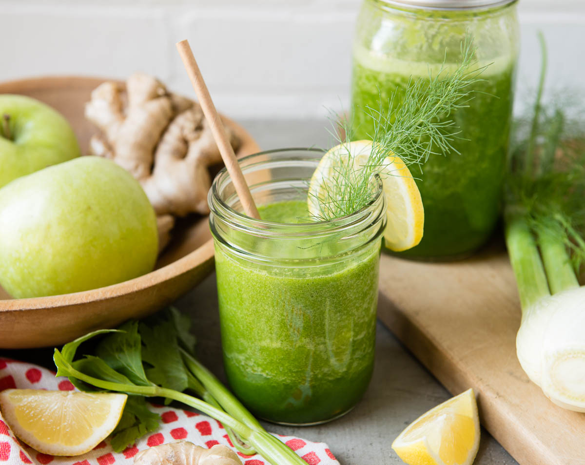 apple celery smoothie in a glass jar with a paper straw surrounded by fresh ingredients.