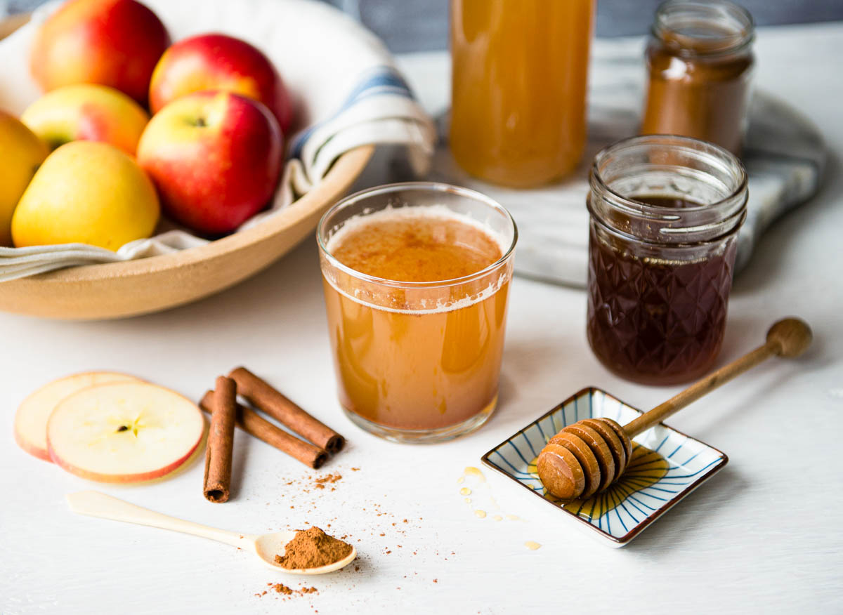 apple cider vinegar drink in a short glass next to a jar of honey, cinnamon sticks, apple slices and a teaspoon of cinnamon.